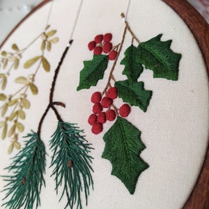 May include: Embroidered hoop art featuring a sprig of holly with red berries, a pine branch, and a sprig of mistletoe. The embroidery is done on a white fabric background.