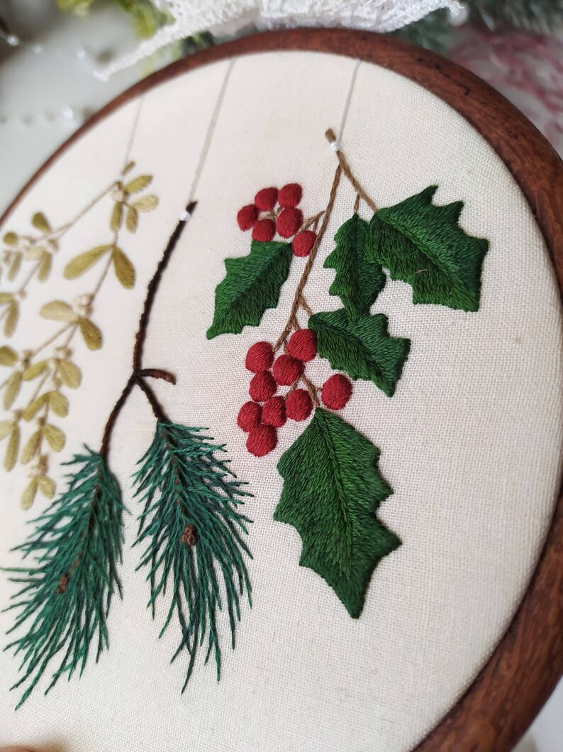 May include: Embroidered hoop art featuring a sprig of holly with red berries, a pine branch, and a sprig of mistletoe. The embroidery is done on a white fabric background.