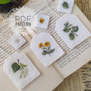 Puede incluir: Etiquetas de tela blanca bordadas con diseños florales, incluyendo girasoles y hojas. Cada etiqueta tiene una cuerda para colgar. Las etiquetas están dispuestas sobre una página de libro vintage.