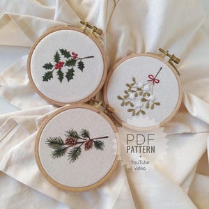Può includere: Tre cerchi da ricamo con diversi motivi a tema natalizio. I cerchi sono in legno e hanno un tessuto bianco teso sopra di essi. I motivi sono un ramo di agrifoglio con bacche rosse, un ramo di vischio con bacche bianche e un ramo di pino con pigne. Il testo "PDF PATTERN + YouTube video" è stampato su un'etichetta bianca.