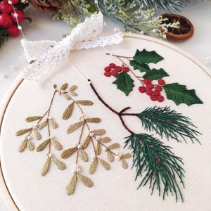May include: A white embroidery hoop with a white lace bow on top. The hoop contains an embroidered design of green pine branches, red holly berries, and green mistletoe.