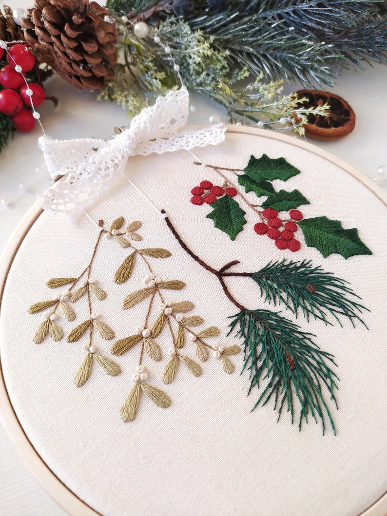 May include: A white embroidery hoop with a white lace bow on top. The hoop contains an embroidered design of green pine branches, red holly berries, and green mistletoe.