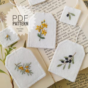 May include: Five white felt tea bag ornaments with embroidered botanical designs including yellow flowers, green leaves, and black olives. Each piece features a white string and a PDF PATTERN + YouTube video label is visible on the left side.
