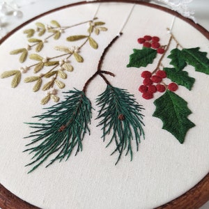 May include: Embroidered hoop art featuring a sprig of mistletoe, holly with red berries, and pine branches with pine cones. The embroidery is done on white fabric and framed in a brown wooden hoop.