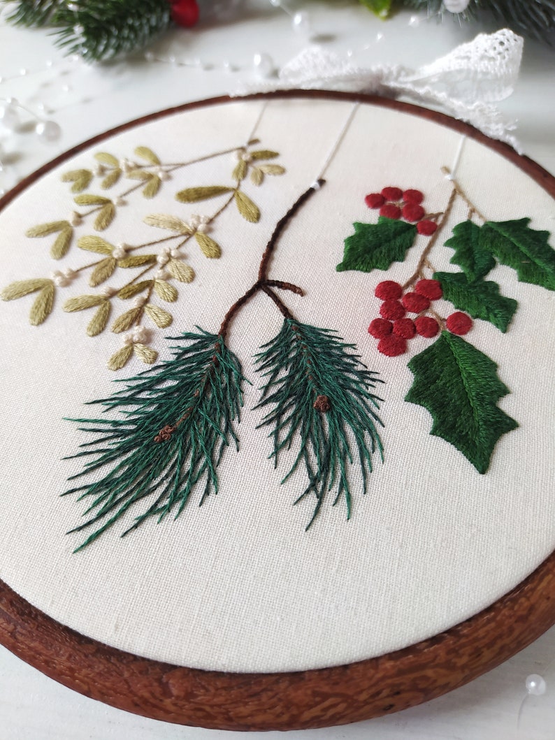 May include: Embroidered hoop art featuring a sprig of mistletoe, holly with red berries, and pine branches with pine cones. The embroidery is done on white fabric and framed in a brown wooden hoop.