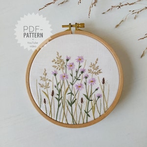 以下が含まれることがあります： 白い背景に紫色の野花と緑色の茎が描かれた花柄の刺繍が施されたフープアート。フープは天然木製です。