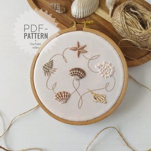 May include: A wooden embroidery hoop with a cream-colored fabric featuring embroidered seashells and a starfish. The hoop is surrounded by a wooden tray with seashells and a ball of twine. The text "PDF - PATTERN + YouTube video" is visible.