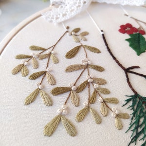 May include: Close-up of a hand-stitched embroidery hoop featuring a sprig of green mistletoe with white berries. The mistletoe is stitched in a delicate, realistic style on a white linen fabric.