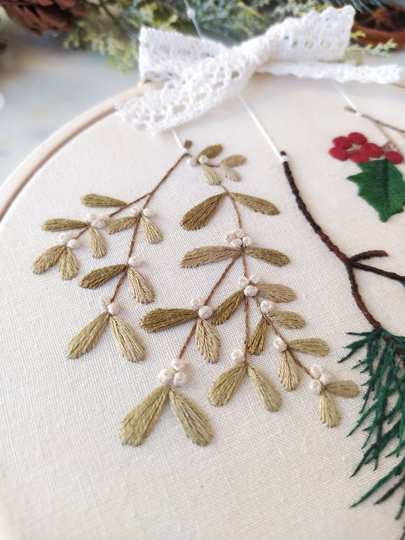 May include: Close-up of a hand-stitched embroidery hoop featuring a sprig of green mistletoe with white berries. The mistletoe is stitched in a delicate, realistic style on a white linen fabric.
