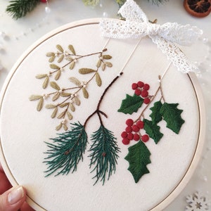 May include: A white embroidery hoop with a green pine branch, red berries, and green leaves with white berries. The hoop is decorated with a white lace bow.