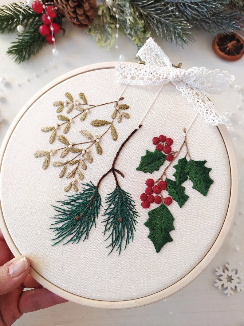 May include: A white embroidery hoop with a green pine branch, red berries, and green leaves with white berries. The hoop is decorated with a white lace bow.
