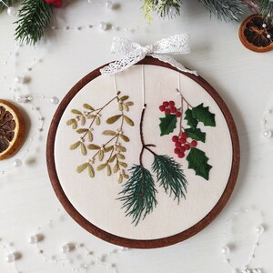 May include: Embroidered hoop art featuring a sprig of mistletoe, holly with red berries, and pine needles on a white background. The hoop is made of wood and has a white lace bow.