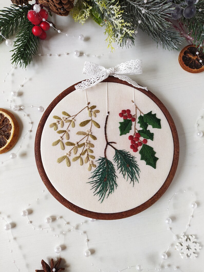 May include: Embroidered hoop art featuring a sprig of mistletoe, holly with red berries, and pine needles on a white background. The hoop is made of wood and has a white lace bow.