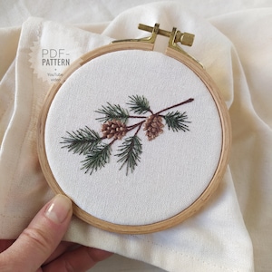 May include: Embroidered pine branch with two pinecones in a wooden embroidery hoop. The embroidery uses green and brown thread on white fabric. The text "PDF-PATTERN" and "YouTube video" is visible.