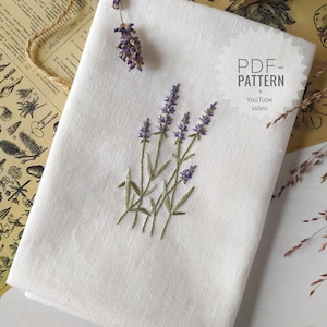 Op de afbeelding: Een gevouwen witte linnen doek geborduurd met takjes lavendel. De lavendel is paars met groene stelen en bladeren. De tekst "PDF-PATTERN + YouTube video" staat in de rechterbovenhoek. Gedroogde lavendel en touw staan op de achtergrond.