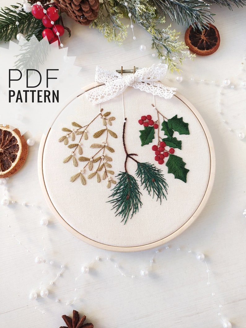 May include: A white embroidery hoop with a design of holly, mistletoe, and pine branches. The design is embroidered in green, red, and gold thread. The hoop is decorated with a white lace bow.