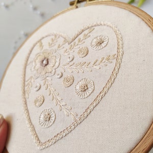 Puede incluir: Un aro de bordado blanco con un dise&ntilde;o en forma de coraz&oacute;n beige con motivos florales y un contorno delicado. El aro es de madera y tiene un acabado natural.