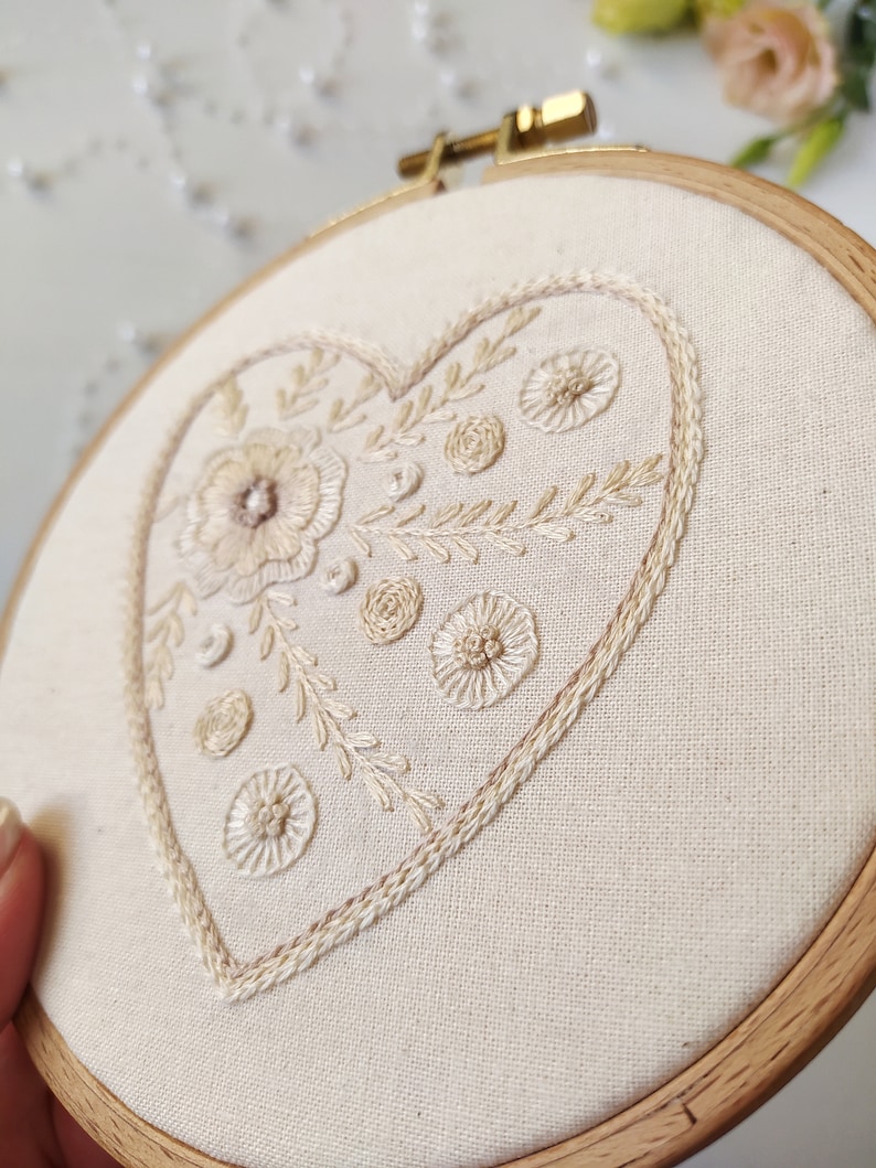 Puede incluir: Un aro de bordado blanco con un dise&ntilde;o en forma de coraz&oacute;n beige con motivos florales y un contorno delicado. El aro es de madera y tiene un acabado natural.
