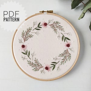 May include: A white embroidery hoop with a floral wreath design. The wreath is made up of green leaves, small white flowers, and larger pink flowers with burgundy centers.