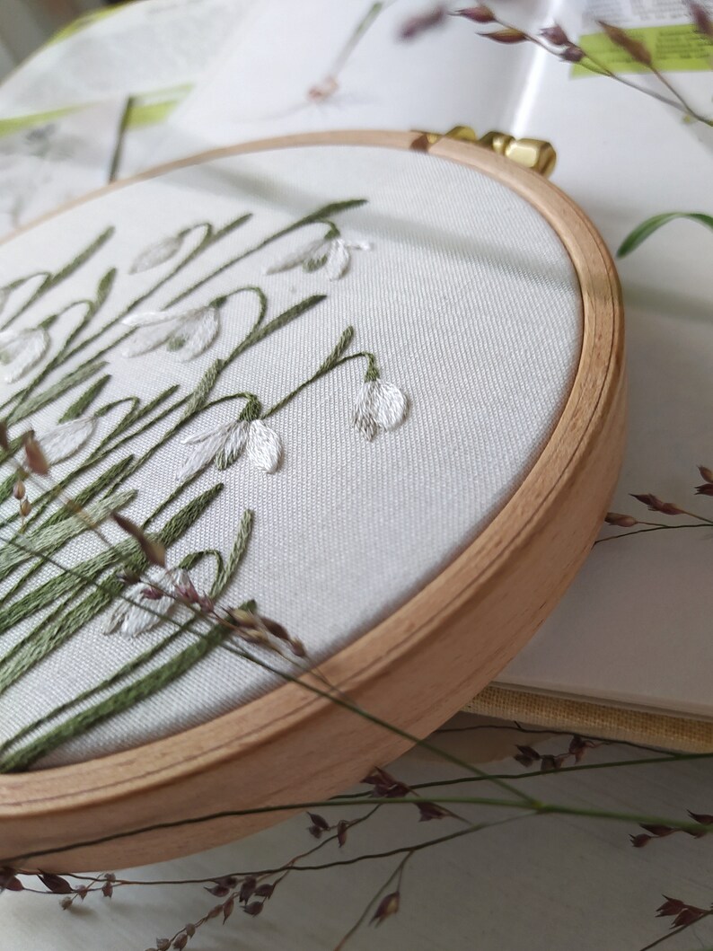 Puede incluir: Primer plano de un aro de bordado blanco con un dise&ntilde;o de hojas verdes y flores blancas. El aro est&aacute; hecho de madera y tiene un acabado natural.