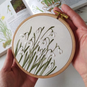Puede incluir: Un aro de bordado blanco con un dise&ntilde;o de campanillas blancas con tallos y hojas verdes. El aro es de madera y tiene un acabado natural.