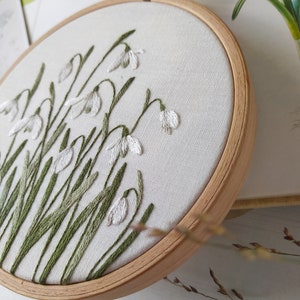 Puede incluir: Un aro de bordado blanco con un dise&ntilde;o de campanillas blancas con tallos verdes sobre un fondo de tela blanca.