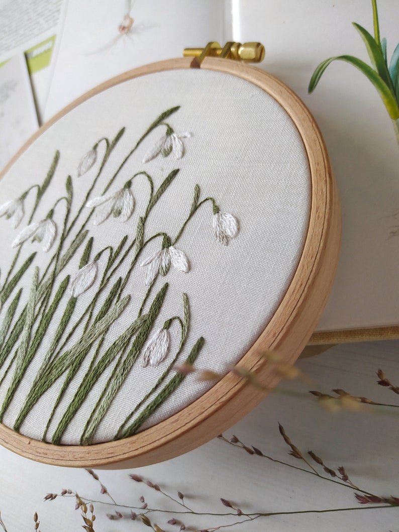 Puede incluir: Un aro de bordado blanco con un dise&ntilde;o de campanillas blancas con tallos verdes sobre un fondo de tela blanca.