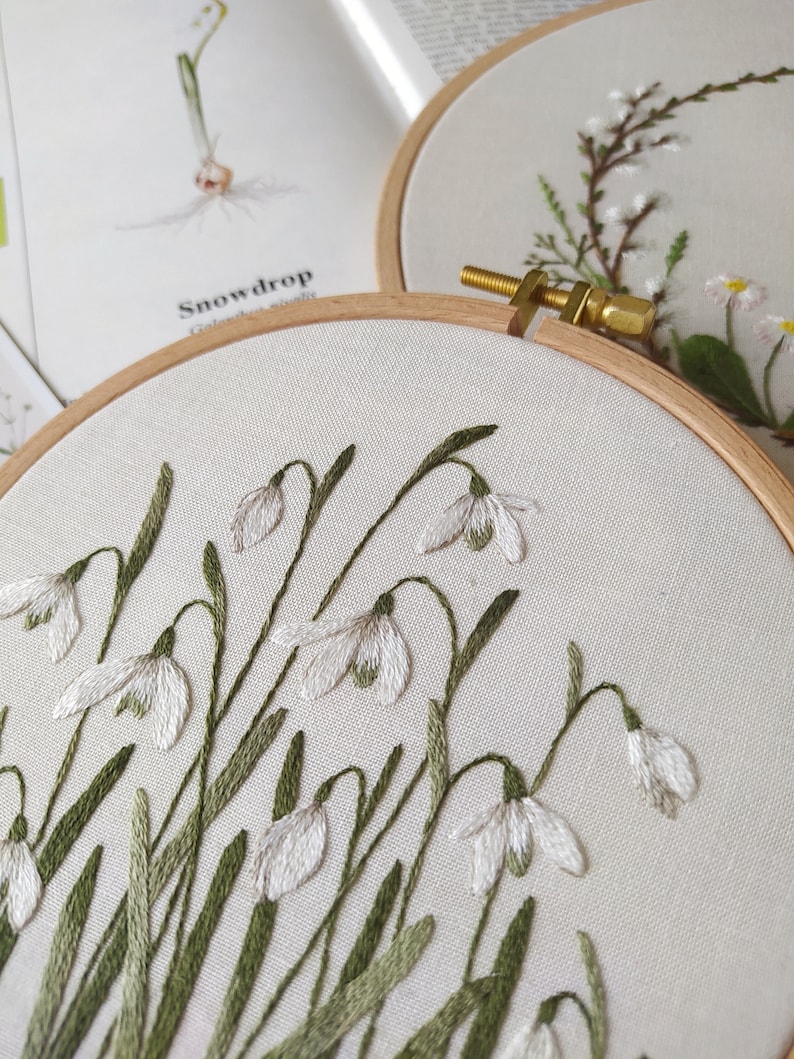 Puede incluir: Primer plano de un aro de bordado blanco con un dise&ntilde;o de flores de campanilla de invierno blancas con tallos y hojas verdes. El bordado est&aacute; sobre un fondo de tela blanca.