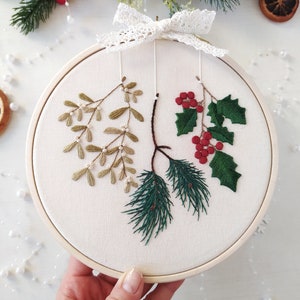 May include: A white embroidery hoop with a green pine branch, a green holly branch with red berries, and a sprig of mistletoe with white beads. The hoop is decorated with a white lace bow.