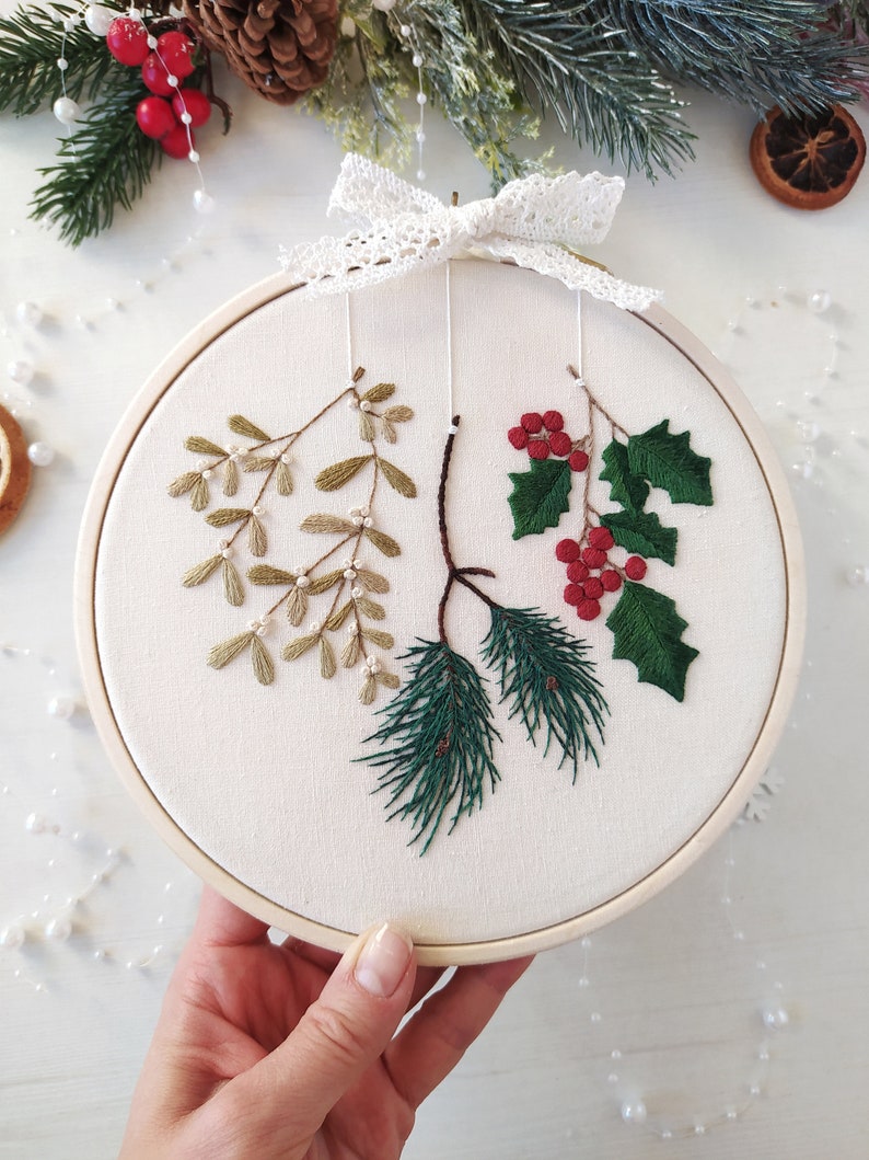 May include: A white embroidery hoop with a green pine branch, a green holly branch with red berries, and a sprig of mistletoe with white beads. The hoop is decorated with a white lace bow.