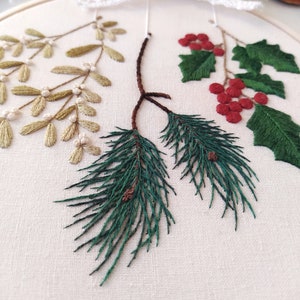May include: A close-up of a white embroidery hoop with three different types of greenery embroidered on it. The greenery includes sprigs of holly with red berries, sprigs of mistletoe with white berries, and pine branches with brown cones.