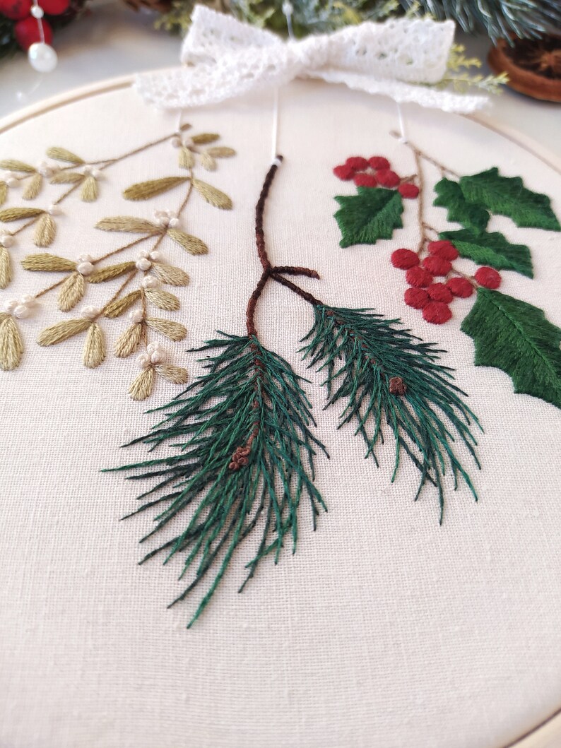 May include: A close-up of a white embroidery hoop with three different types of greenery embroidered on it. The greenery includes sprigs of holly with red berries, sprigs of mistletoe with white berries, and pine branches with brown cones.