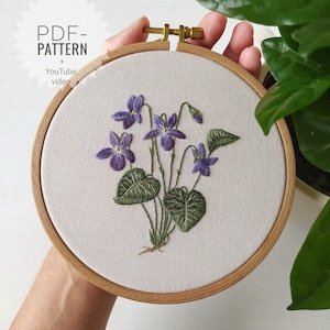 May include: Embroidered hoop art featuring a cluster of purple violets with green leaves. The hoop is made of wood and has a white fabric background. The text "PDF-PATTERN + YouTube video" is visible in the top left corner.