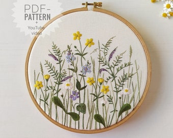 May include: Embroidered hoop art featuring a floral design with yellow, purple, and white flowers and green leaves. The hoop is made of wood and has a white fabric background. The text "PDF-PATTERN + YouTube video" is visible in the upper left corner of the image.