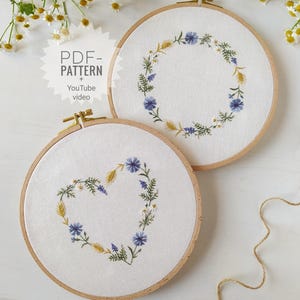 Può includere: Due telai da ricamo con disegni floreali. Uno presenta una ghirlanda a forma di cuore, l'altro una ghirlanda circolare. Entrambi hanno ricami floreali blu, gialli e bianchi su tessuto bianco, incorniciati in telai di legno. Il testo "PDF - PATTERN + YouTube video" è visibile.