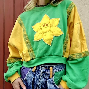 Può includere: Una felpa verde e gialla con un'ampia applicazione di narciso giallo. Le maniche sono gialle e i polsini sono a coste. La felpa è abbinata a jeans in denim lavato acido a vita alta con dettagli di tasche viola.