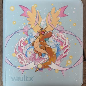 Peut inclure: Un classeur de cartes à collectionner bleu clair orné d'une créature orange ailée au centre, entourée de rubans roses, de petites étoiles jaunes et de silhouettes bleues d'autres créatures. Le nom de marque vaultx est imprimé en bas à gauche.
