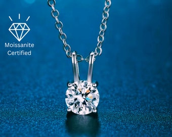May include: A silver pendant necklace with a round, sparkling moissanite gemstone. The necklace has a delicate chain and a simple, elegant design. The image includes the text "Moissanite Certified".