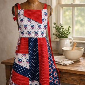May include: A patchwork apron featuring a patriotic color scheme of red, white, and blue. The fabric displays patterns of stars, polka dots, and eagle emblems. It includes a square bib with a red triangular accent and two white buttons at the waist.