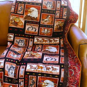 Può includere: Una trapunta con design patchwork e pannelli rettangolari che mostrano immagini di aquile calve, lupi, cavalli in corsa, ceramiche e figure stilizzate. La tavolozza dei colori include tonalità di marrone, nero, arancione e bianco con bordo rosso.