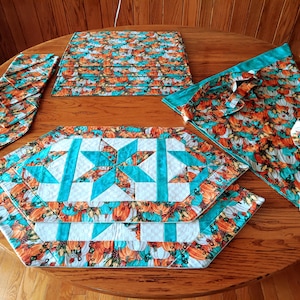 May include: A set of quilted fabric table linens featuring a pumpkin print in orange, white, and teal. The collection includes rectangular placemats, a folded table runner, and a large triangular cloth, all with a star-patterned center.