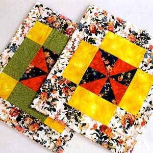 May include: Two rectangular fabric placemats featuring a central pinwheel block pattern in orange and dark blue, surrounded by bright yellow borders and an outer frame of white fabric with a colorful floral print of roses and small blue flowers.