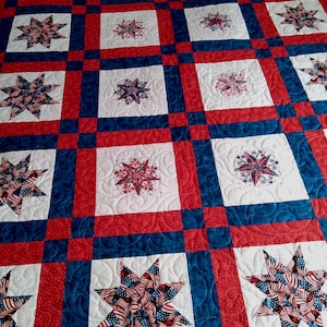 Puede incluir: Una colcha con un patrón de cuadrícula de cuadrados blancos, cada uno con un motivo de estrella, separados por tiras de tela roja y azul. Las estrellas están hechas de tela con patrones rojos, blancos y azules, con un acolchado intrincado.