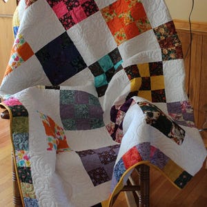 May include: A white quilt with a yellow border and a patchwork design featuring various colors and patterns. The quilt is draped over a wooden rocking chair.