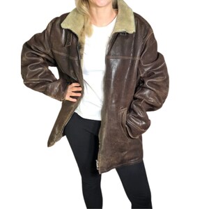 May include: A brown leather jacket with a beige shearling collar. The jacket has a zip closure, two front pockets, and a distressed appearance. The jacket is worn over a white t-shirt and black pants.