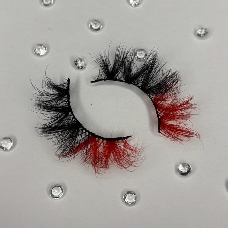 May include: A pair of dramatic false eyelashes with a black base and black lashes, accented with vibrant red tips. The lashes are displayed against a white background with scattered clear rhinestones, creating a glamorous aesthetic.