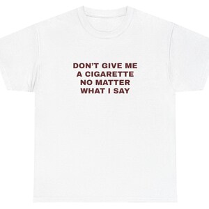 Op de afbeelding: Wit t-shirt met de donkerrode tekst "DON'T GIVE ME A CIGARETTE NO MATTER WHAT I SAY". Het shirt heeft een ronde hals en korte mouwen.