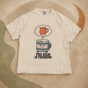 Könnte beinhalten: Ein beiges T-Shirt mit kurzen Ärmeln und einer Grafik eines Cartoon-Brennofens mit traurigem Gesicht, der zu einer Gedankenblase mit einer orangefarbenen Tasse aufschaut. Darunter steht der Text THE KILN IS THINKING in schwarzen Buchstaben.