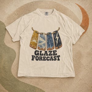 Peut inclure: Un t-shirt beige à manches courtes orné d'un motif représentant quatre étiquettes rectangulaires suspendues à une corde. Les étiquettes illustrent un soleil, des nuages, de la pluie et un éclair. Le texte GLAZE FORECAST est imprimé en noir.