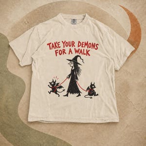 May include: A beige short-sleeve t-shirt featuring a black graphic of a witch walking two small winged demons on red leashes. The red text above the illustration reads, "TAKE YOUR DEMONS FOR A WALK." The shirt has a crew neckline and a relaxed fit.